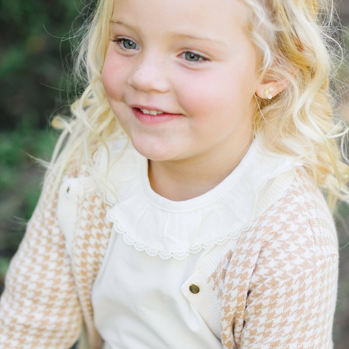Young girl with blonde hair wearing a beige cardigan and white shirt outdoors.