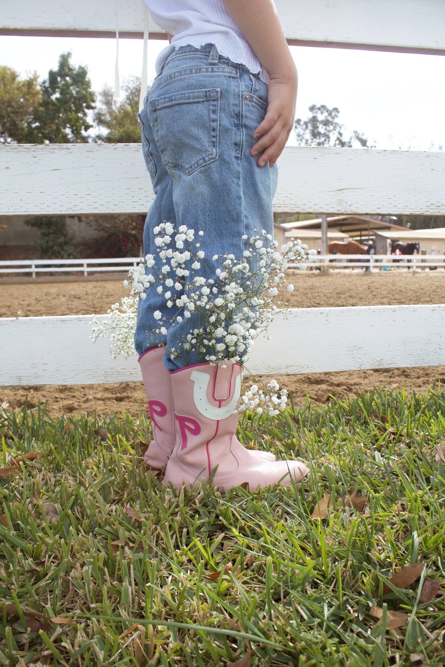 Child wearing pink rain boots with floral decorations, standing on grass with a white fence and trees in the background.