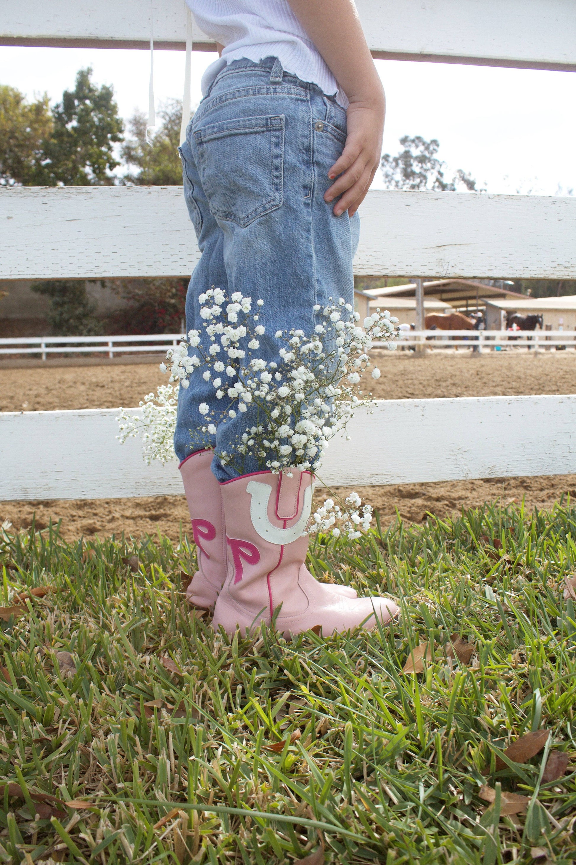 Child wearing pink rain boots with floral decorations, standing on grass with a white fence and trees in the background.
