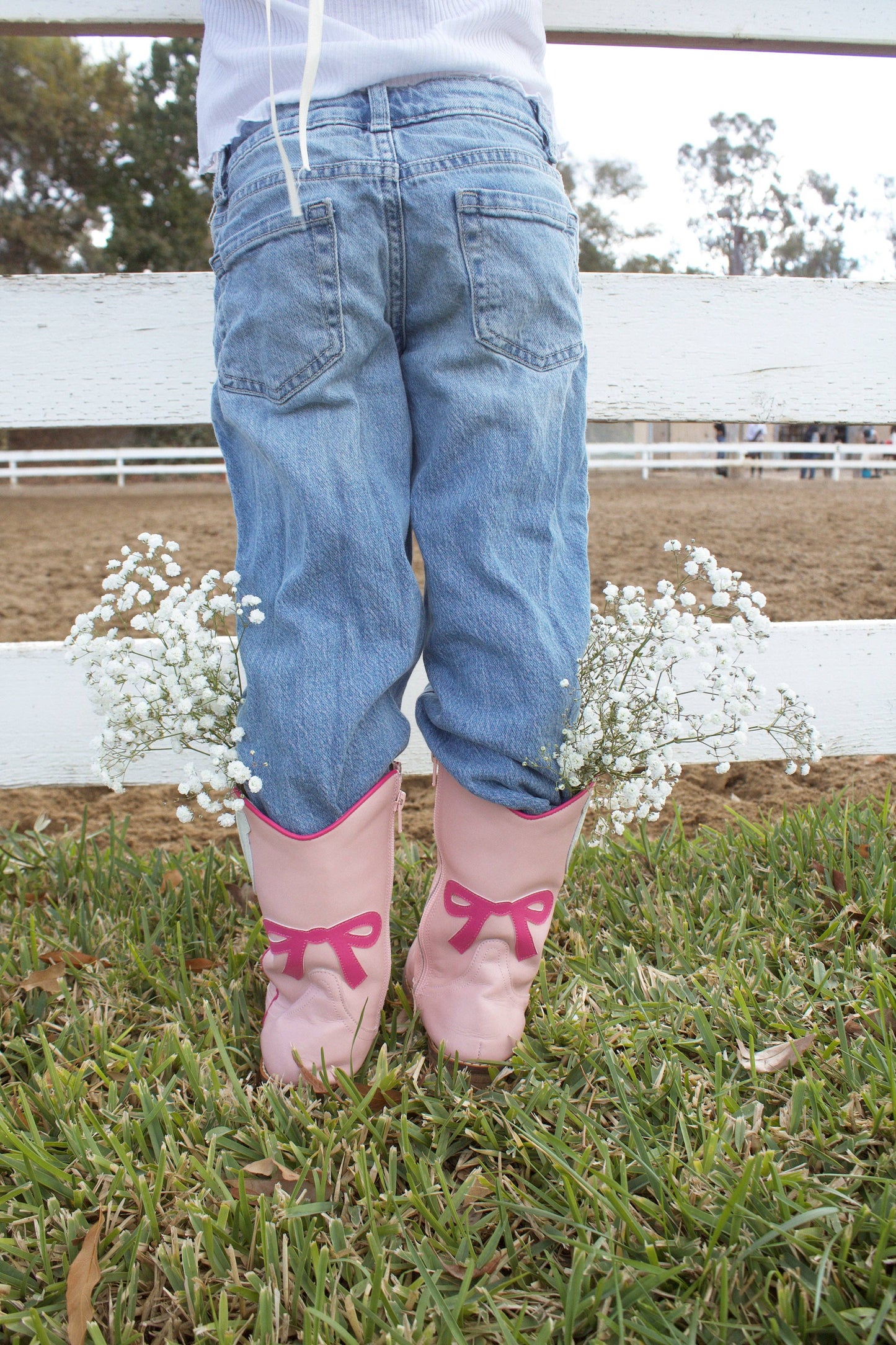 Person wearing blue jeans and pink boots with bows in a grassy area with a white fence and flowers in the background.