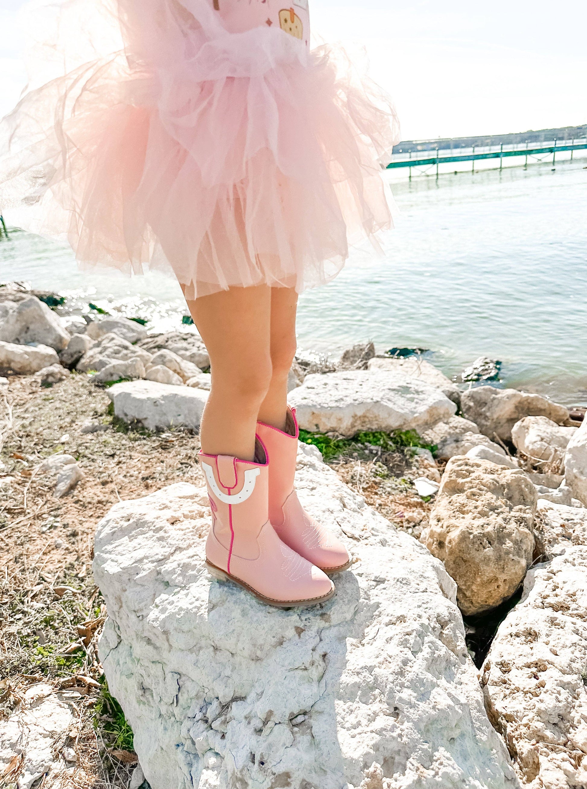 Child wearing pink rain boots standing on rocks by a body of water.