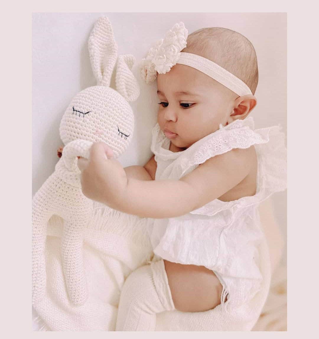 Baby in a white outfit holding a white knit bunny toy on a light background