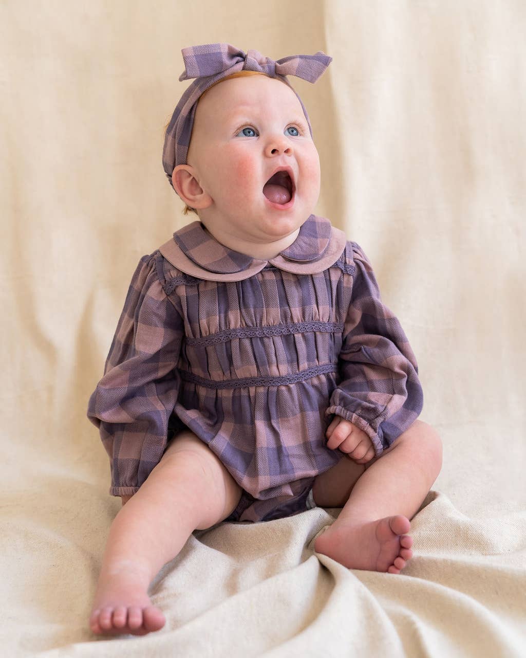 Baby wearing a plaid outfit with a bow headband on a beige background
