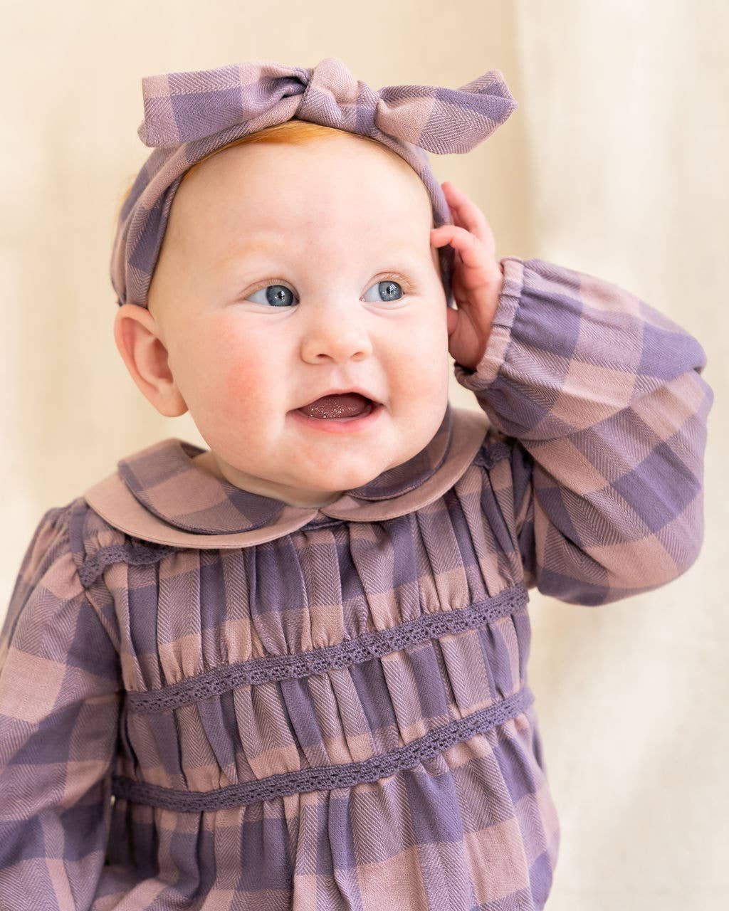 Baby wearing a purple checkered dress and headband against a beige background