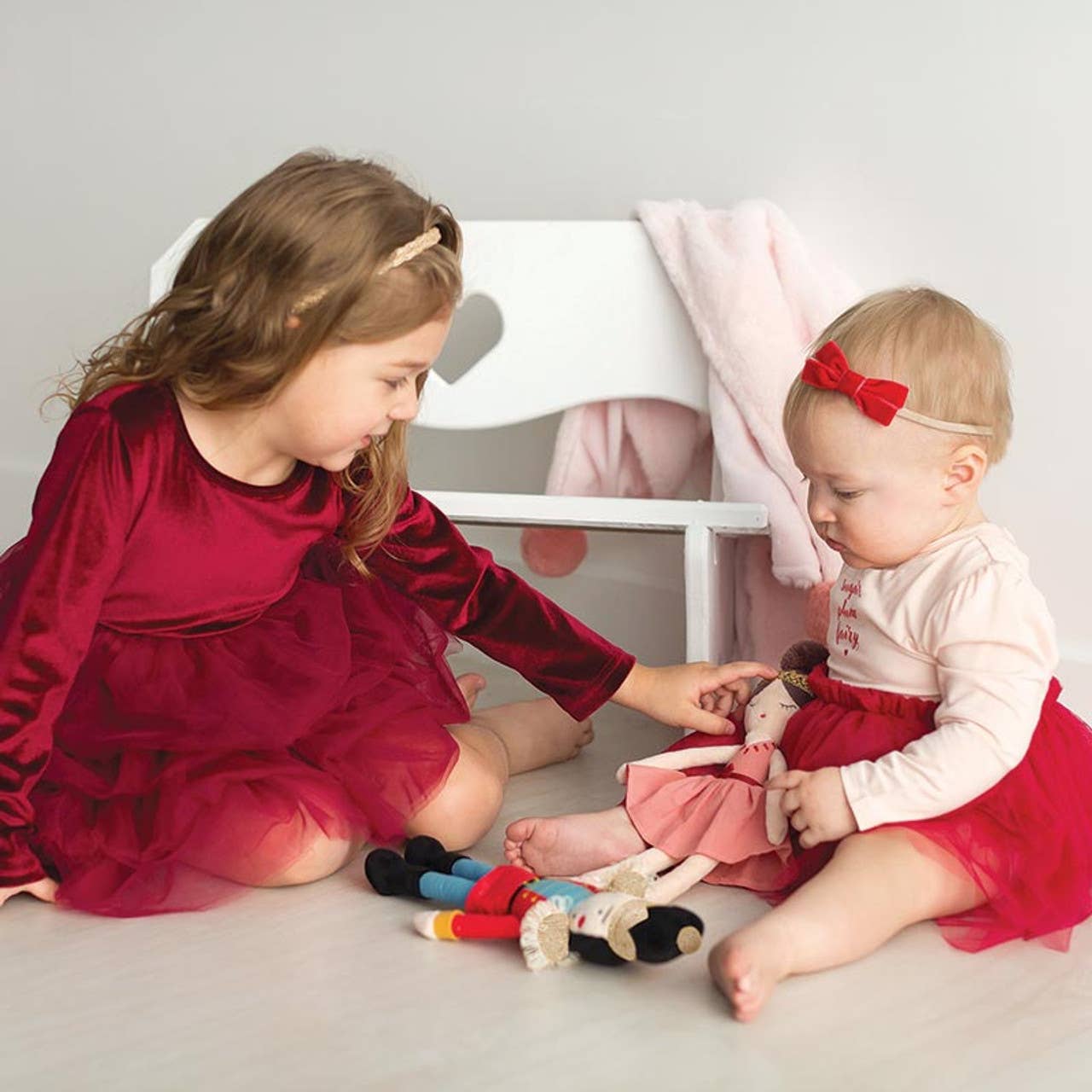 Two young girls in matching red outfits playing with dolls on a light gray floor.