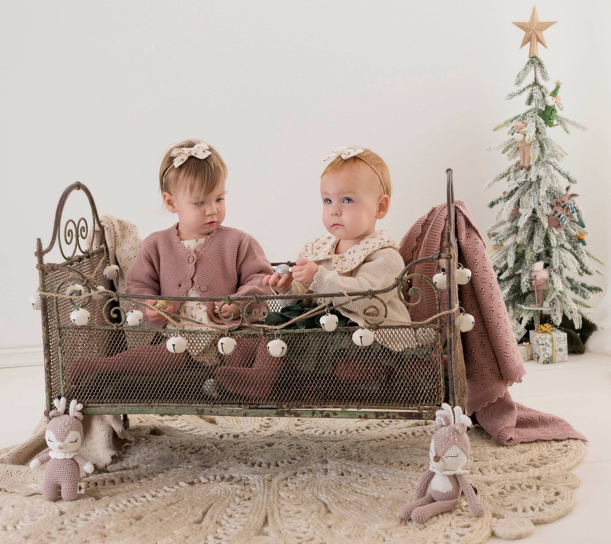 Two young children sitting on a decorative bed with a small Christmas tree in the background.