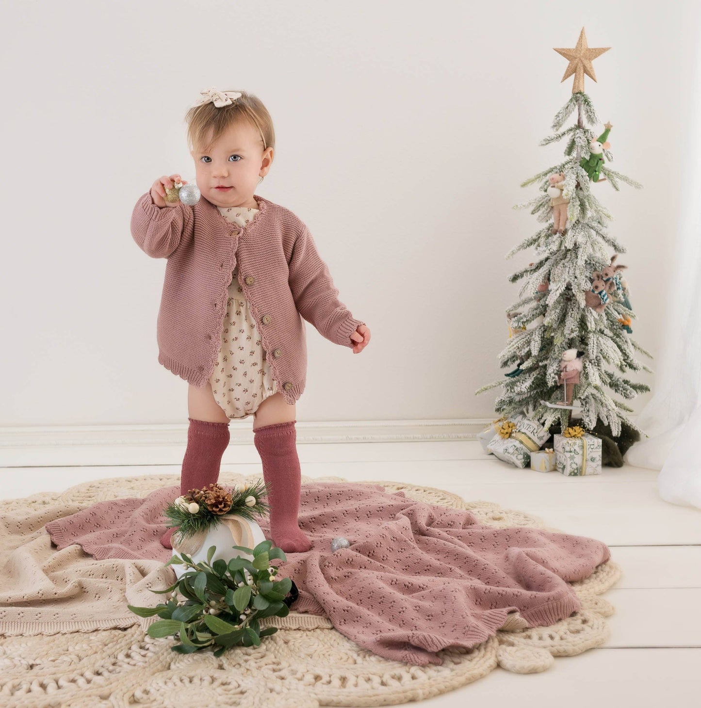 Baby in a pink outfit standing next to a small decorated Christmas tree.
