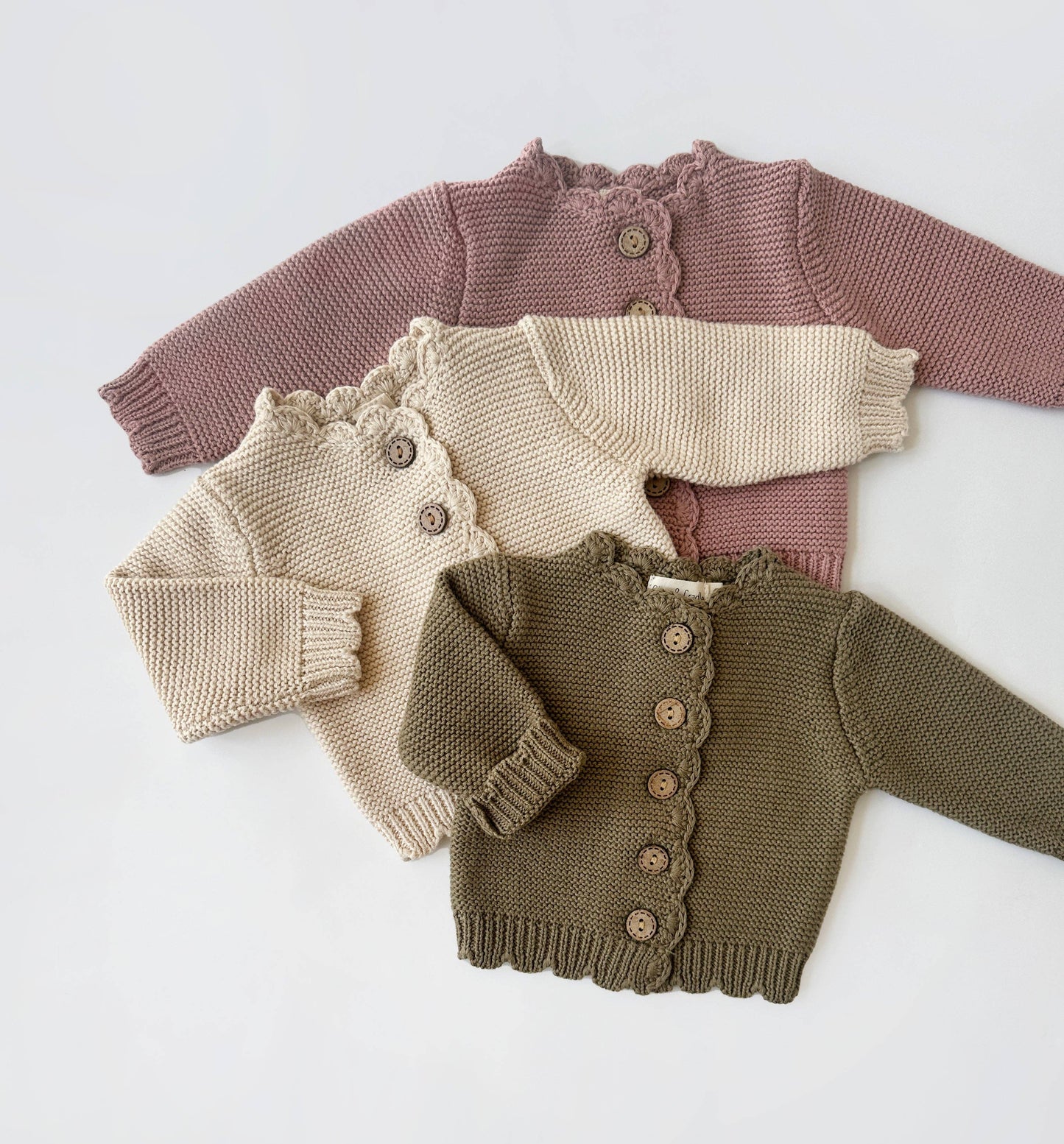 Three knitted baby sweaters in pink, beige, and brown on a white background