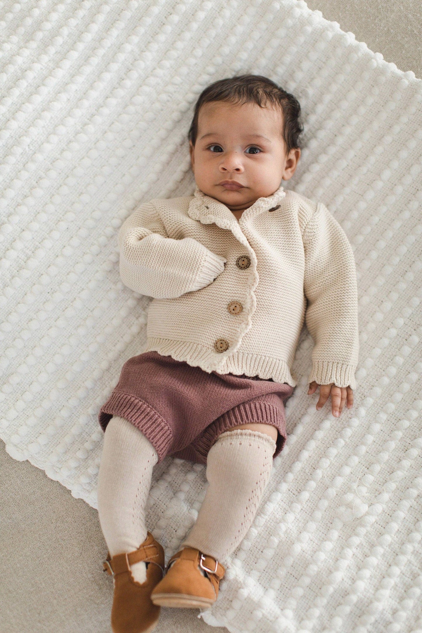 Baby wearing a cream cardigan with buttons, pink shorts, and brown shoes on a textured white blanket.