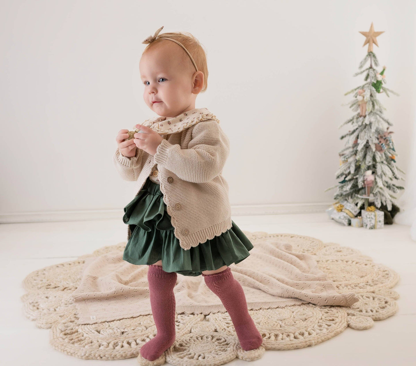 Baby in a beige sweater and green skirt standing on a white floor with a Christmas tree in the background.