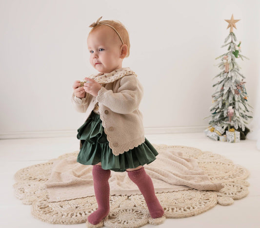 Baby in a beige sweater and green skirt standing on a white floor with a Christmas tree in the background.