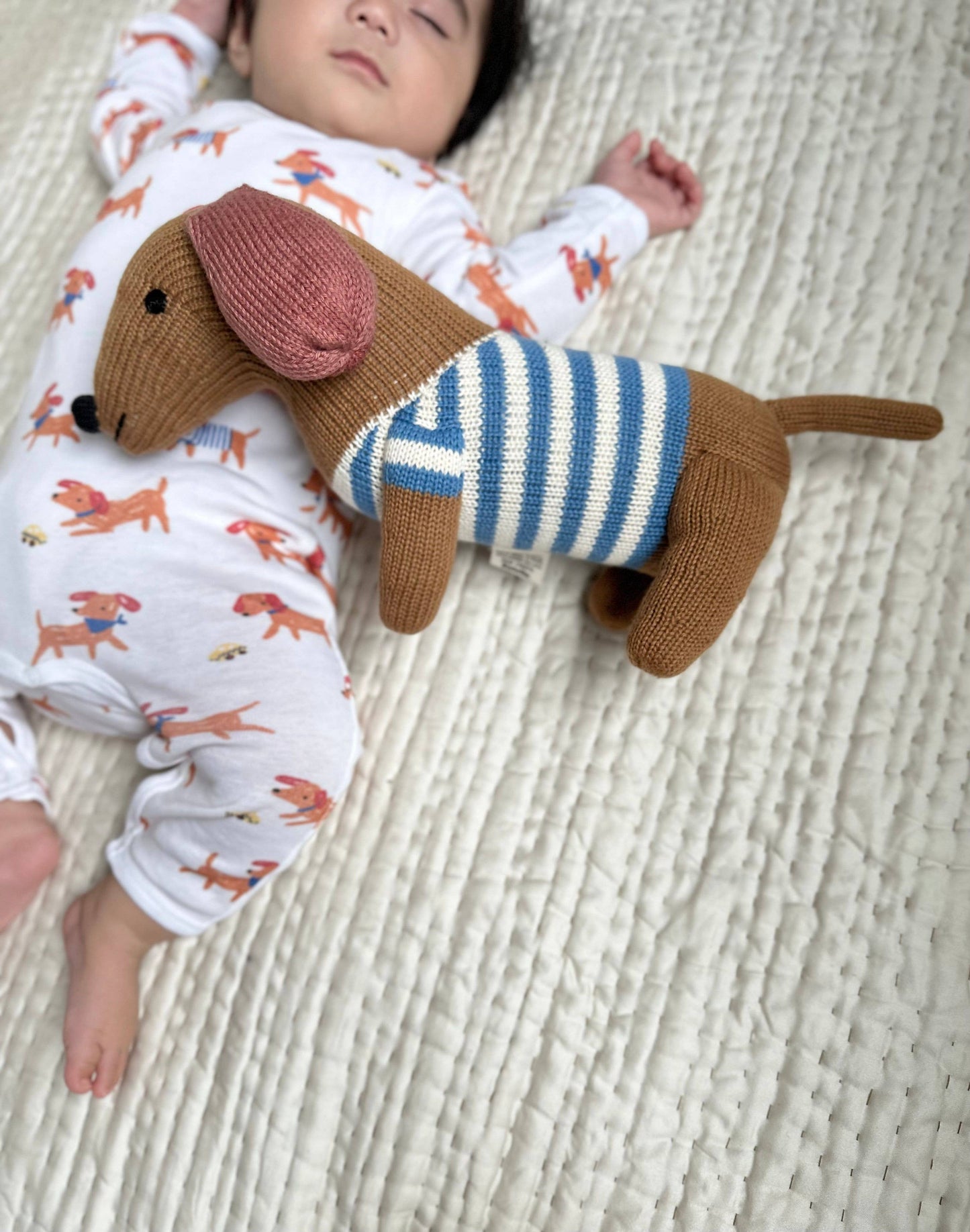 Baby lying on a textured surface with a knitted dachshund toy wearing a striped sweater.