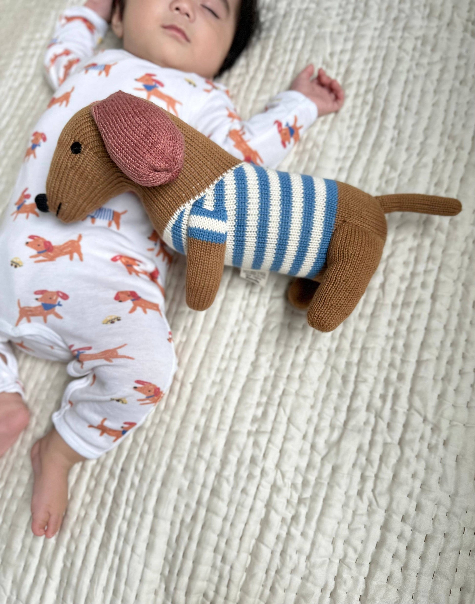 Baby lying on a textured surface with a knitted dachshund toy wearing a striped sweater.