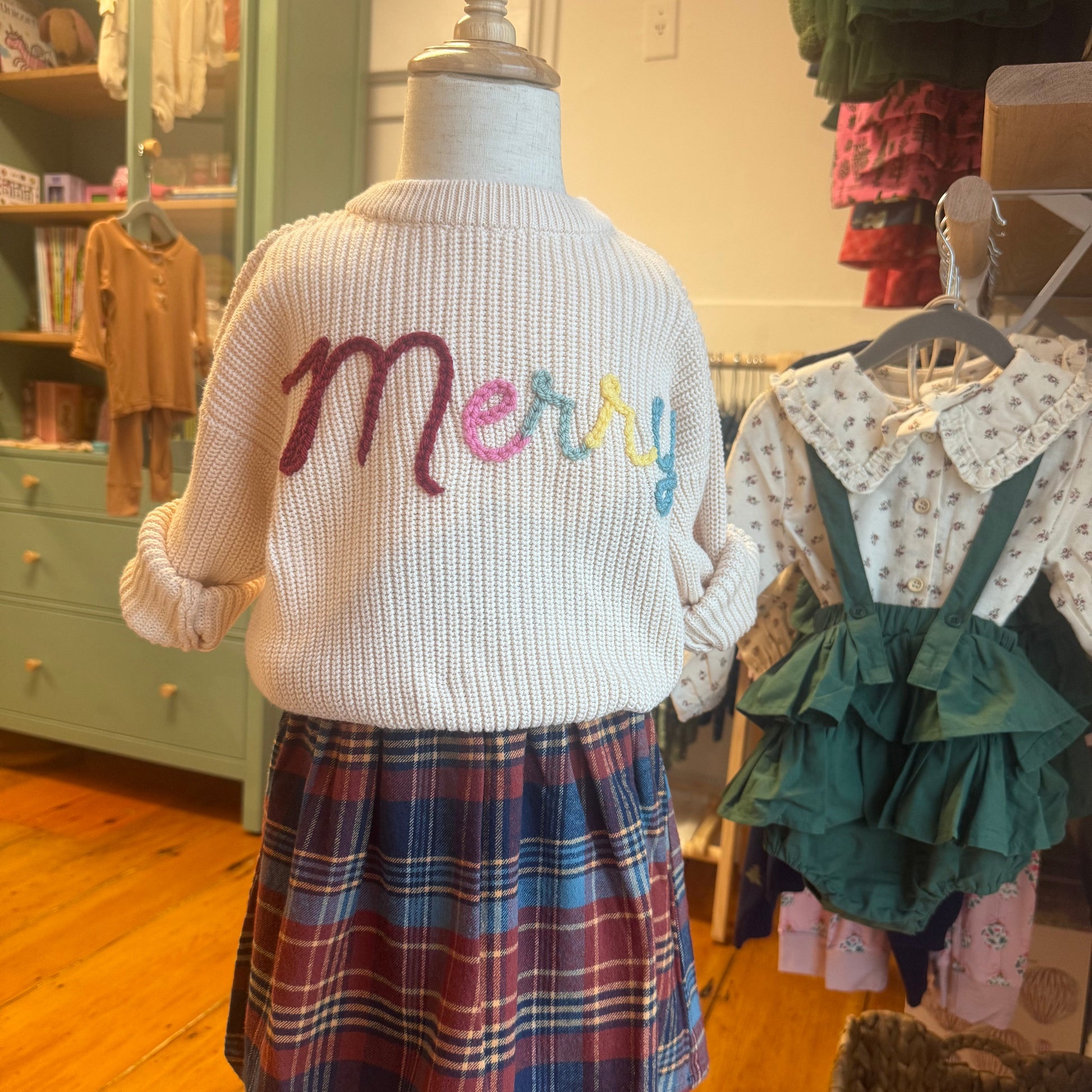Mannequin wearing an apricot sweater with colorful "Merry" stitching and a plaid skirt.