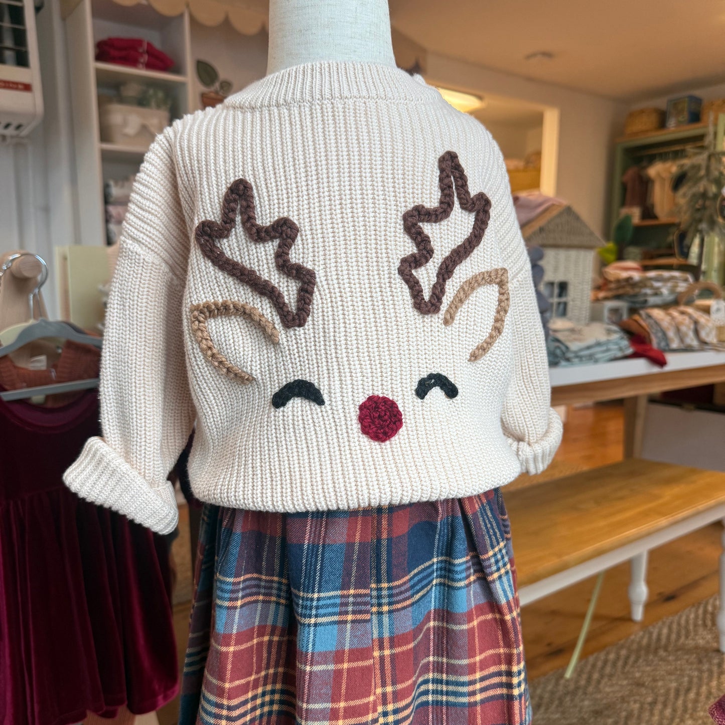 Knit smiling reindeer sweater and plaid skirt on a mannequin in a store setting.