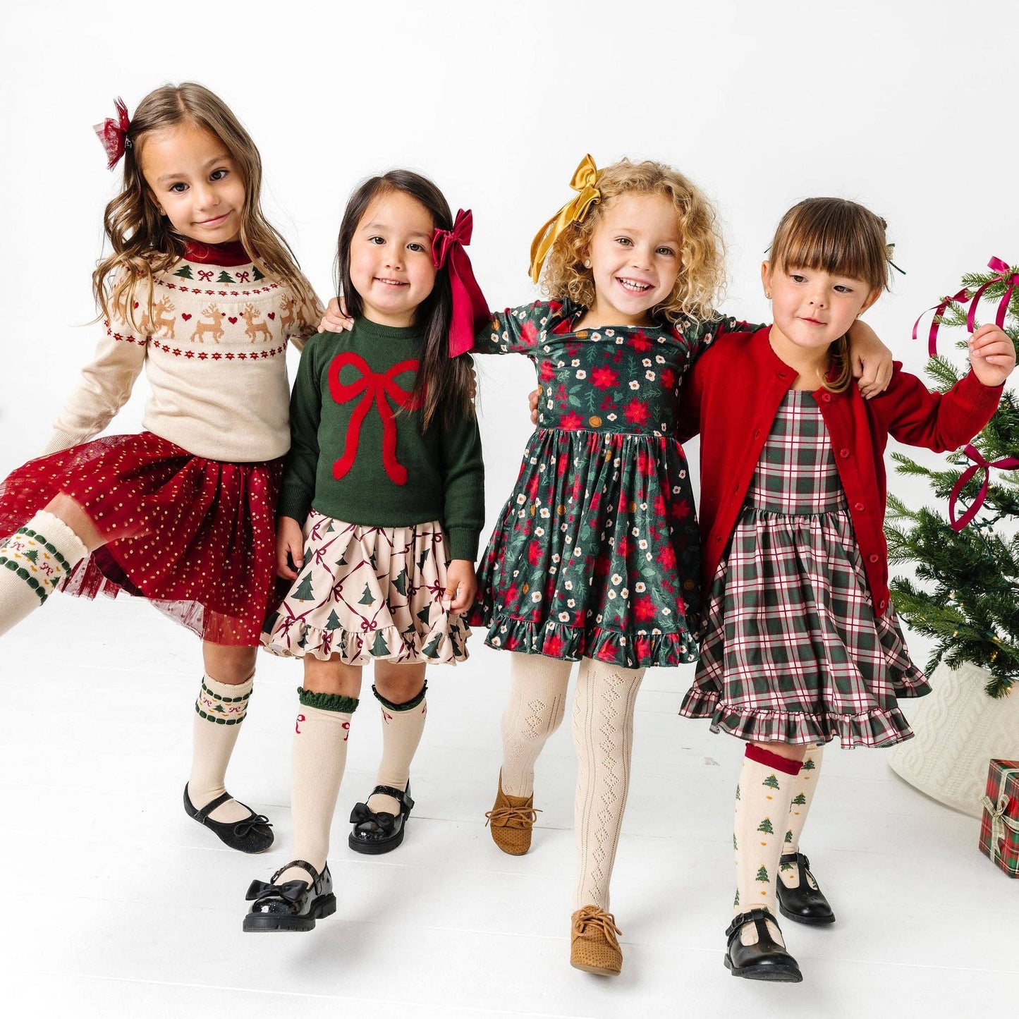 Four young girls in festive outfits standing together with a Christmas tree in the background.