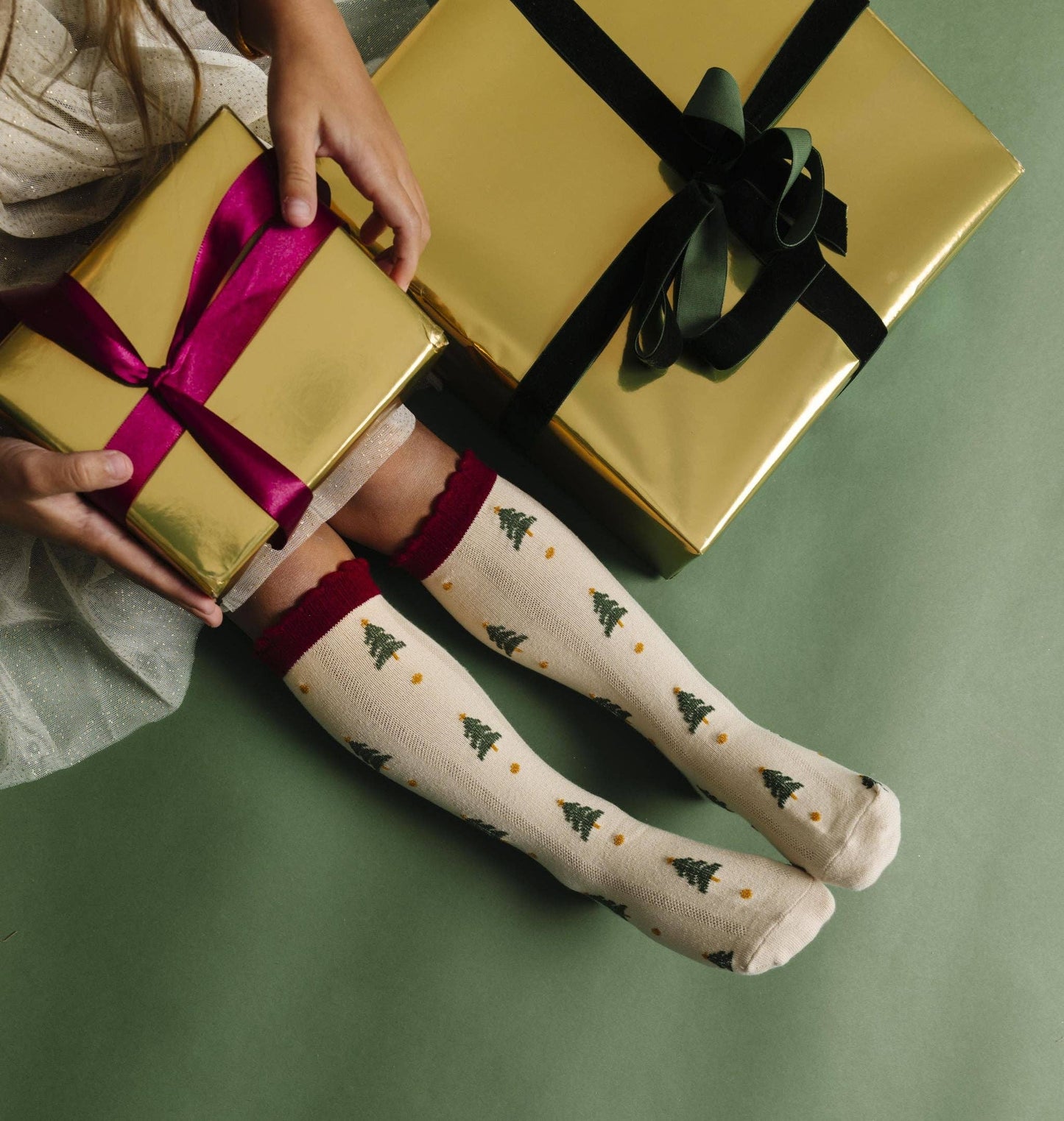 Person holding a gold gift box with a pink ribbon, wearing knee-high socks with tree patterns on a green background.