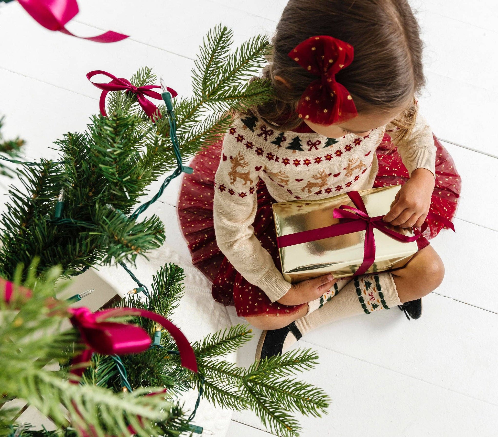 Child in a festive sweater holding a gift next to a Christmas tree with red ribbons.