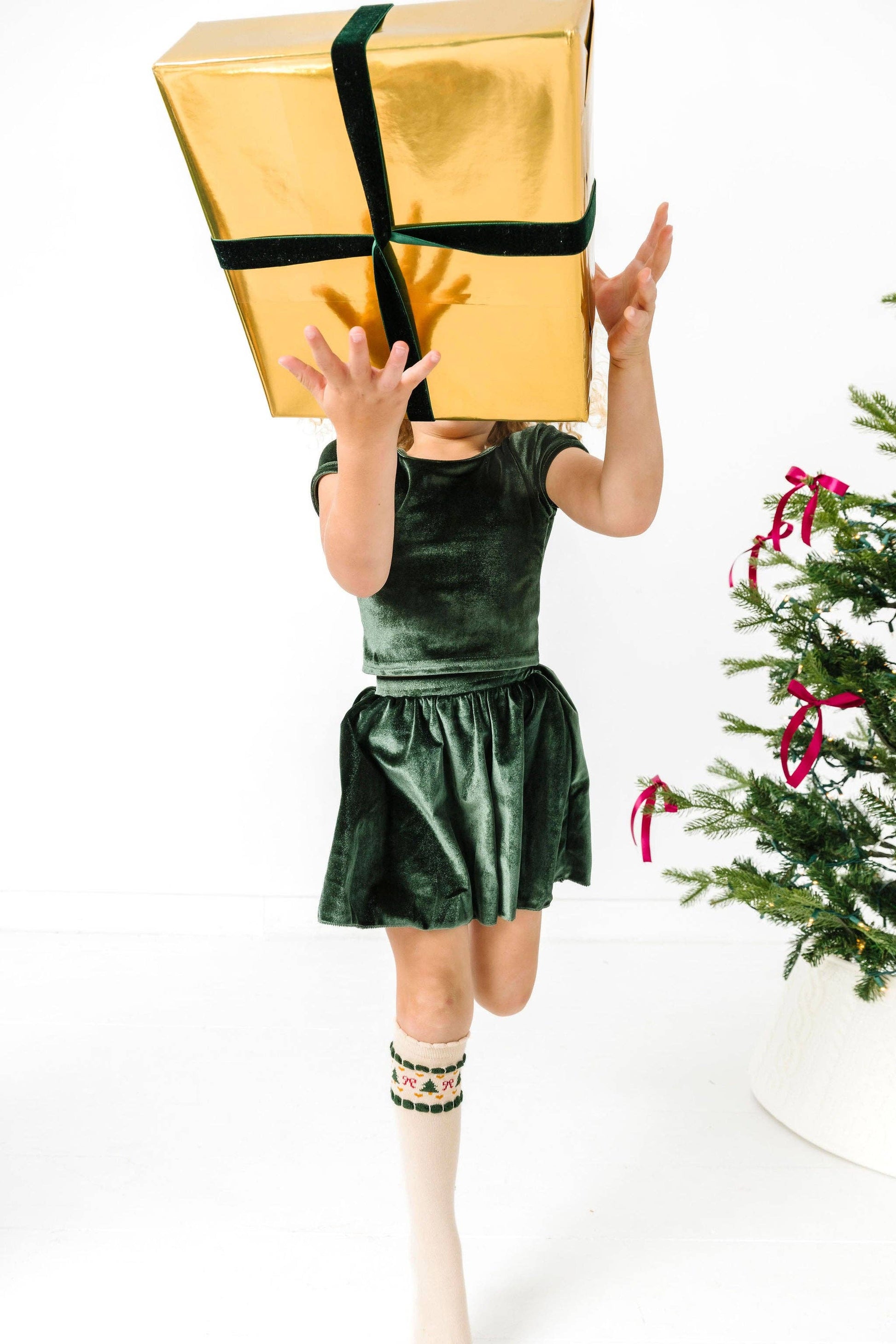 Child in a green dress holding a large gold gift box with a Christmas tree in the background