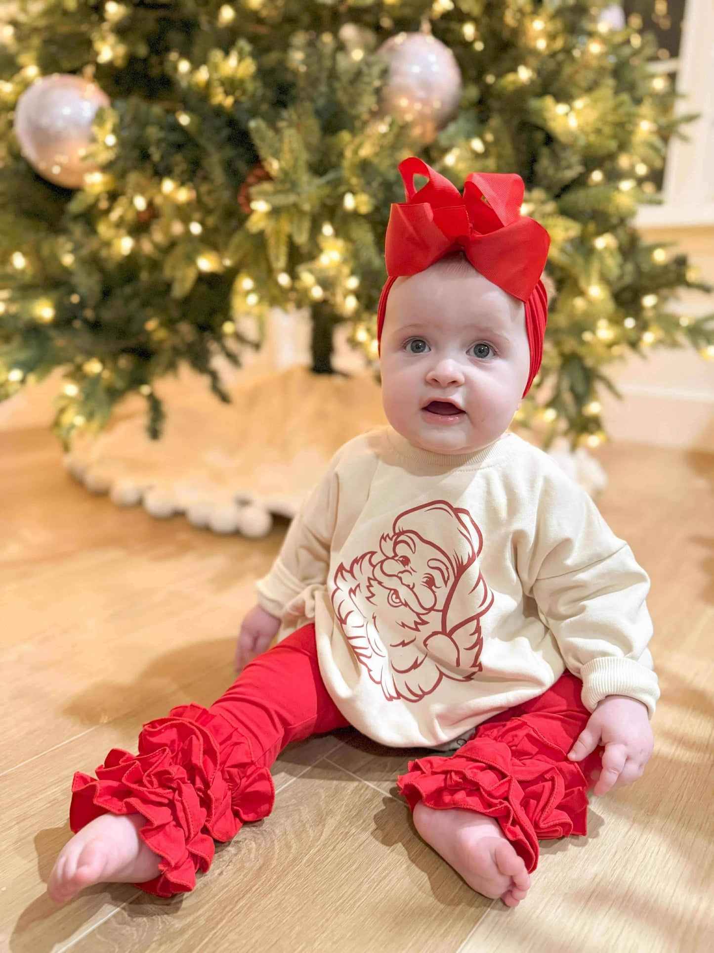 Baby in festive outfit with red bow and ruffled pants sitting in front of a decorated Christmas tree.
