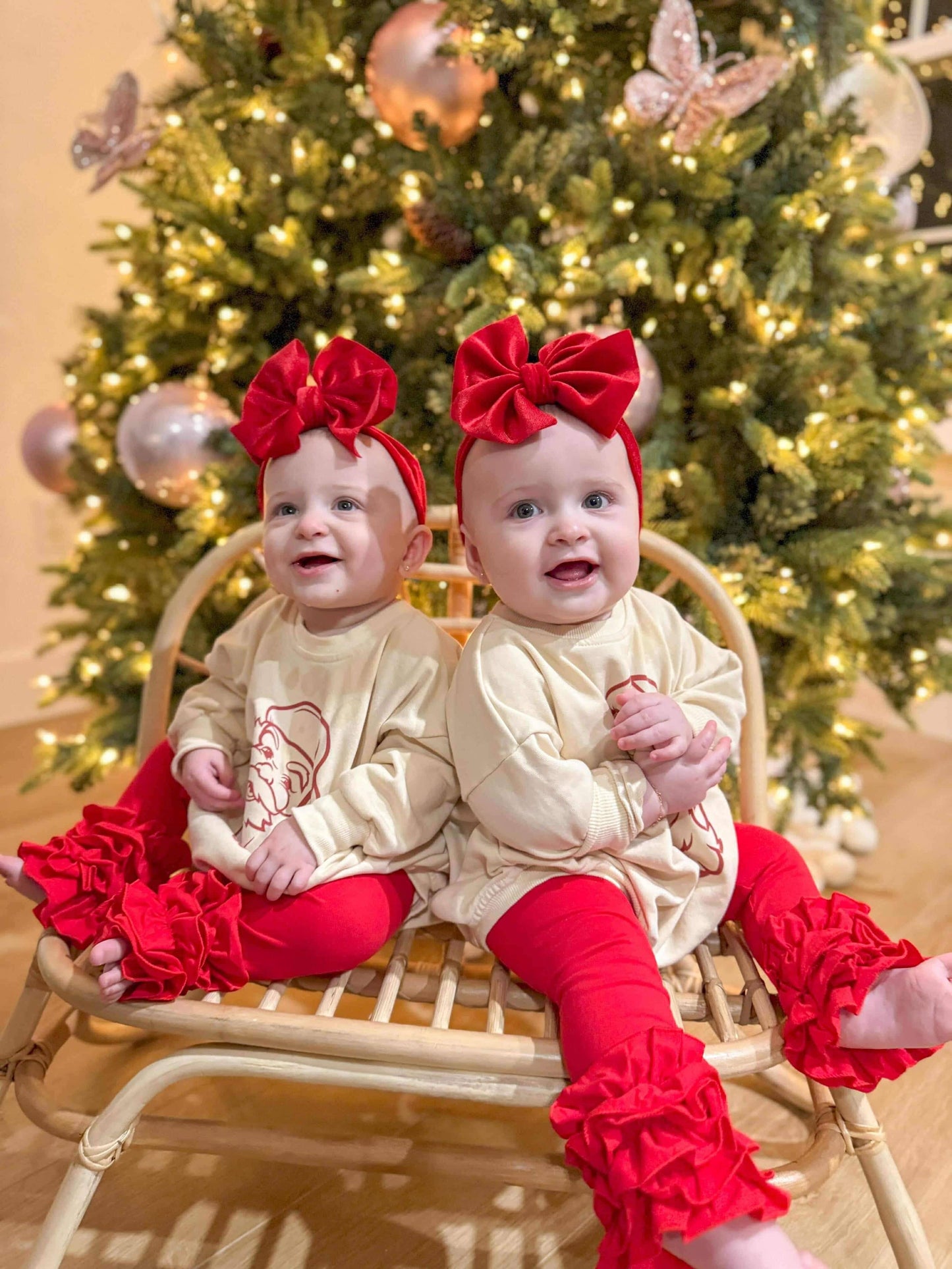 Two babies in matching outfits with red bows and leggings sitting in front of a decorated Christmas tree.