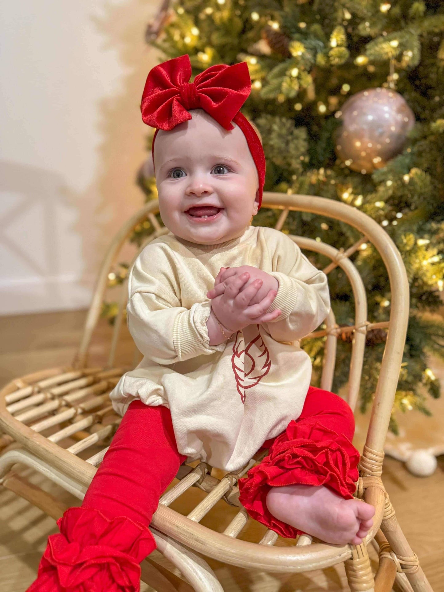 Baby in festive outfit with red bow and leggings sitting in front of a decorated Christmas tree.