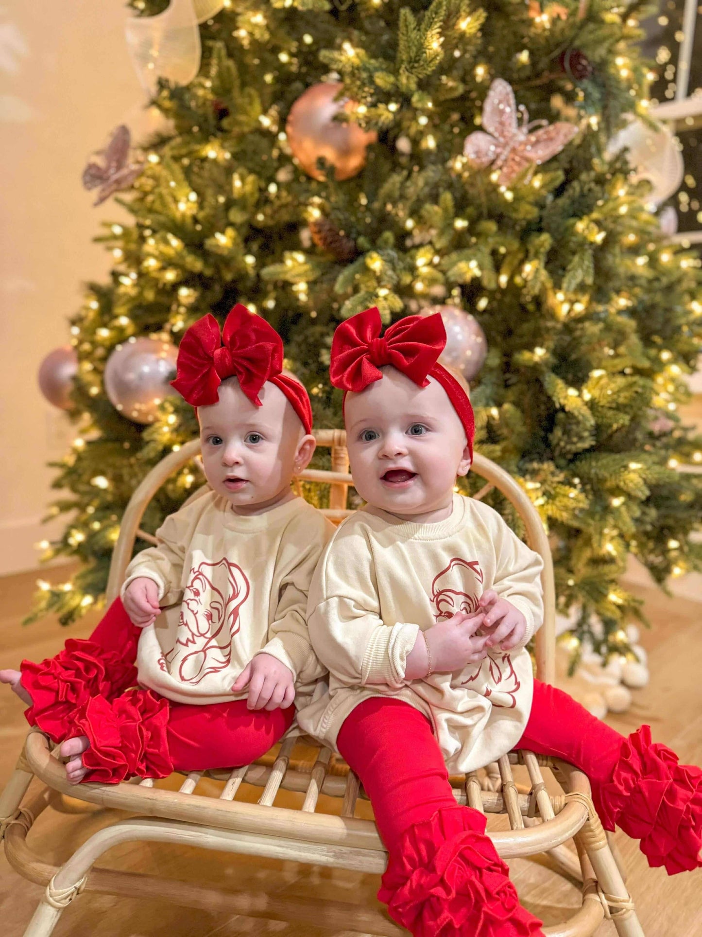 Two children in matching outfits with red bows and ruffled pants sitting in front of a decorated Christmas tree.