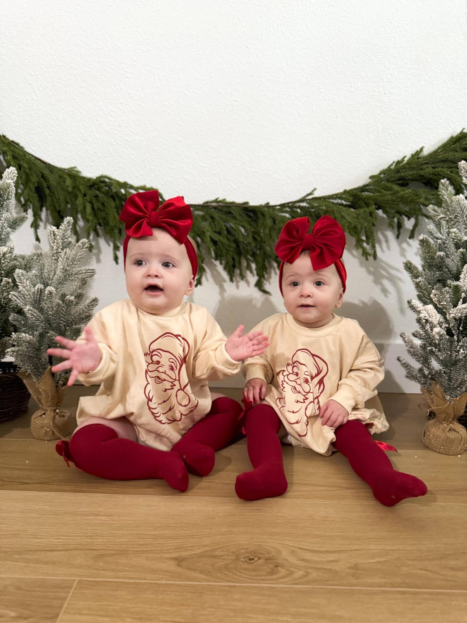 Two babies in matching outfits with red bows and stockings sitting on a wooden floor with Christmas decorations.