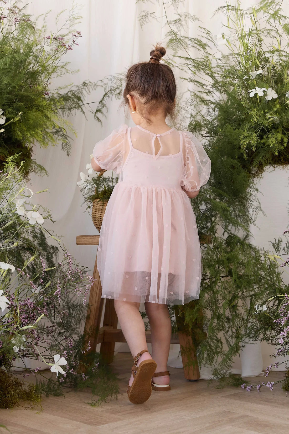 A young girl viewed from behind wearing a light pink tulle dress with sheer puff sleeves, standing on a wooden stool in a lush, green floral indoor setting.