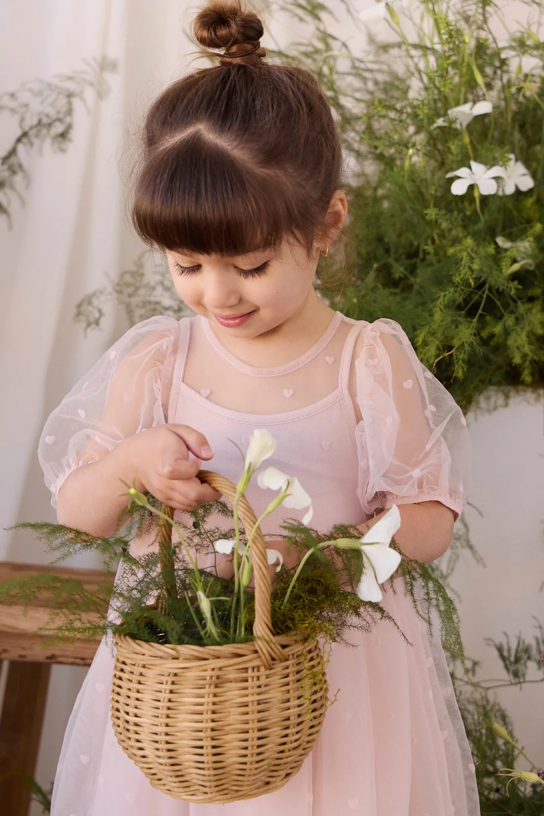 A young girl with brown hair in a bun wearing a light pink dress, holding a wicker basket filled with white flowers and greenery.