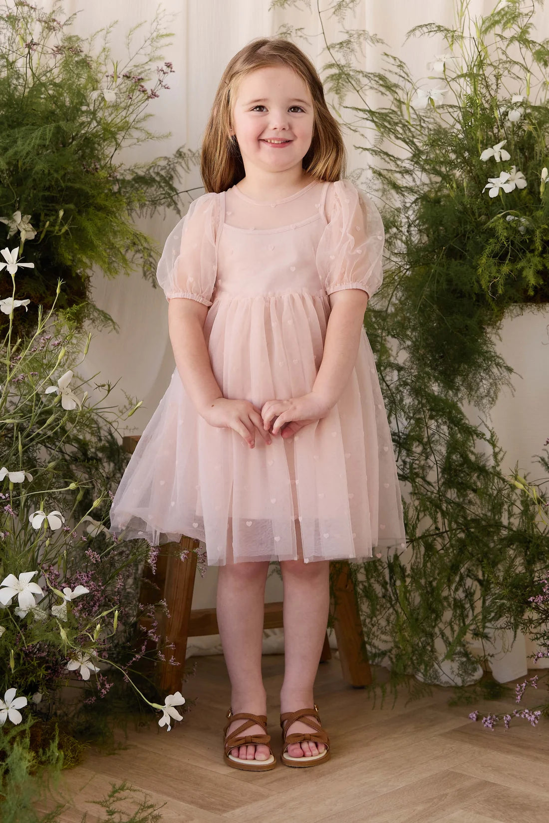 A young girl with light brown hair stands smiling in a dusty pink tulle dress with puff sleeves, styled with brown leather sandals. She is posed in front of a backdrop of green foliage and white flowers.