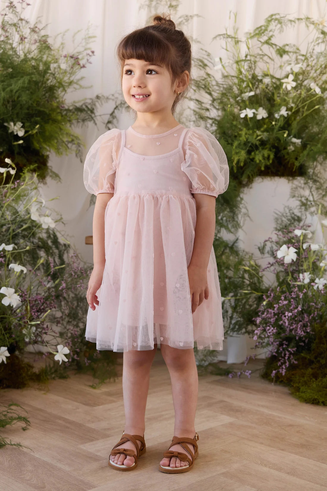 A young girl with her hair in a bun stands wearing a light pink tulle dress with puff sleeves and brown sandals, set against a background of green foliage and white flowers.