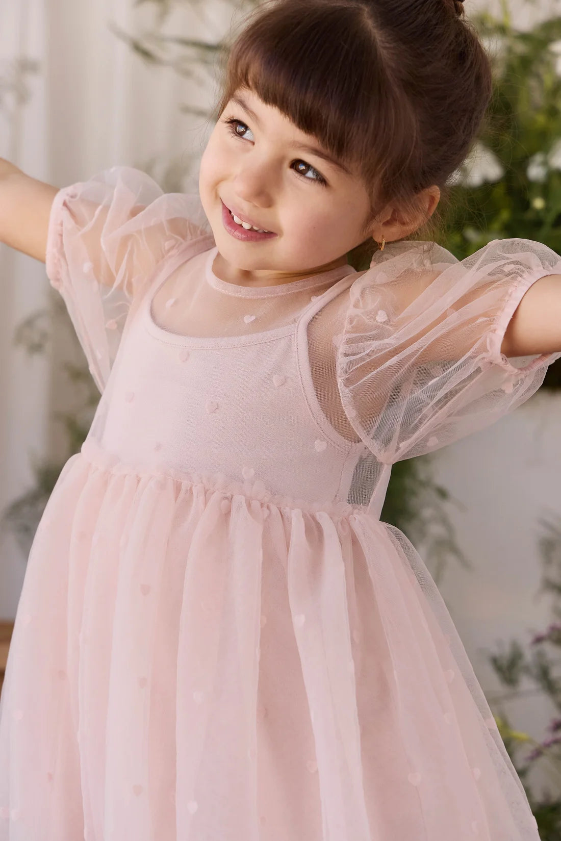 A young girl with her hair in a bun wearing a light pink tulle dress with sheer puff sleeves, smiling and posing with her arms outstretched in a bright, soft-focus indoor setting.