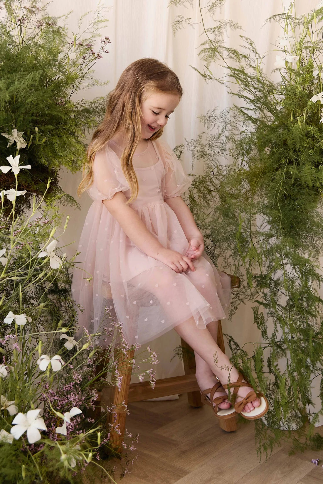 A young girl with long blonde hair sits on a wooden stool in a lush, floral setting, wearing a light pink tulle dress with white embroidered details and brown strappy sandals.
