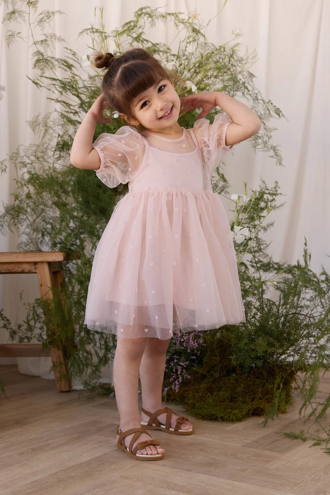 A young girl with her hair in a bun poses in a light pink tulle dress with puffy sleeves and brown strappy sandals, standing in a styled indoor setting with green plants and a wooden stool.