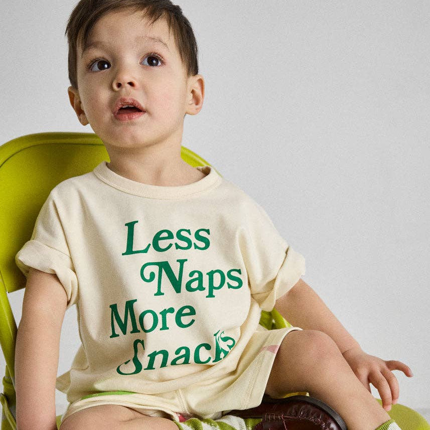 Child wearing a shirt with 'Less Naps More Snacks' text, sitting on a green chair.