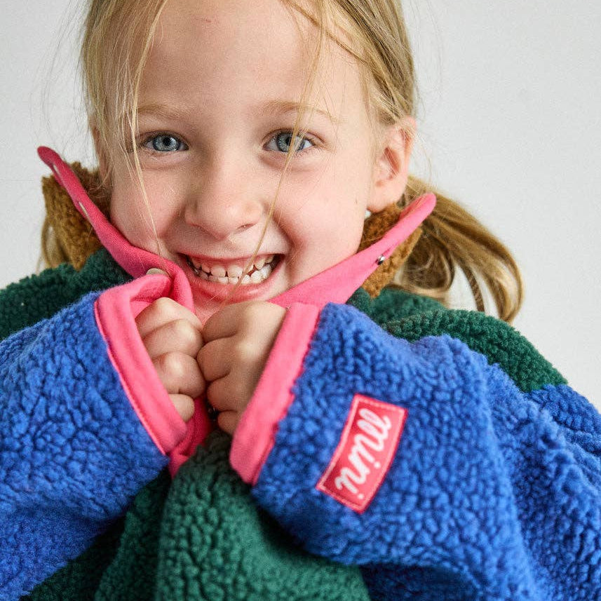 A smiling young girl wearing a fleece jacket with a blue front, green back, and bright pink collar and zipper trim. A red patch on the sleeve shows the brand name 'Milly'.