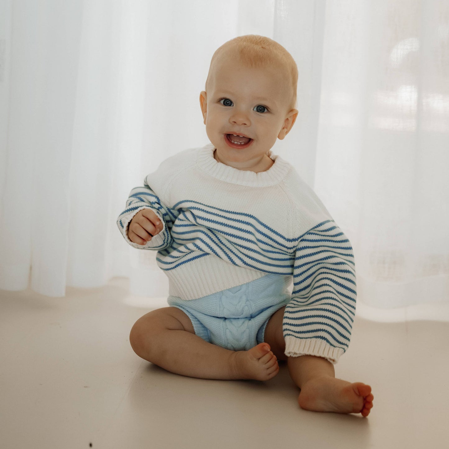 Baby sitting on a white floor wearing a striped sweater and blue shorts with a white curtain background.