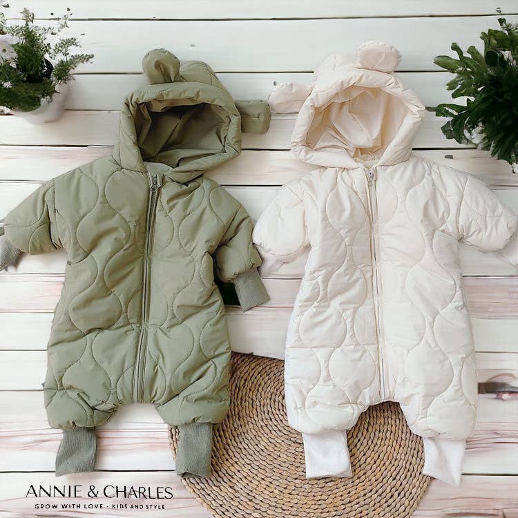 Two baby snowsuits, one green and one white, on a wooden floor with a plant and rug in the background. Brand 'Annie & Charles' visible.