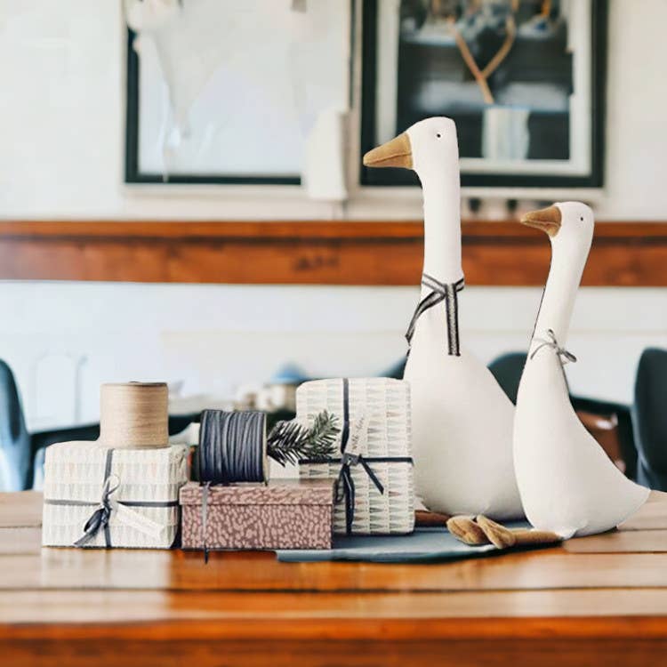 Two white goose figurines on a wooden surface with decorative items in the background.
