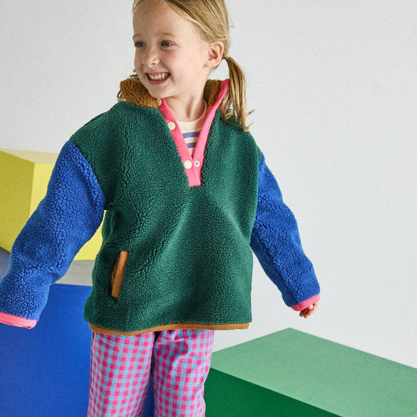 A young girl smiling and posing in a colorful fleece pullover sweater and pink-and-white gingham pajama pants, standing on a pink block in front of large geometric color blocks.