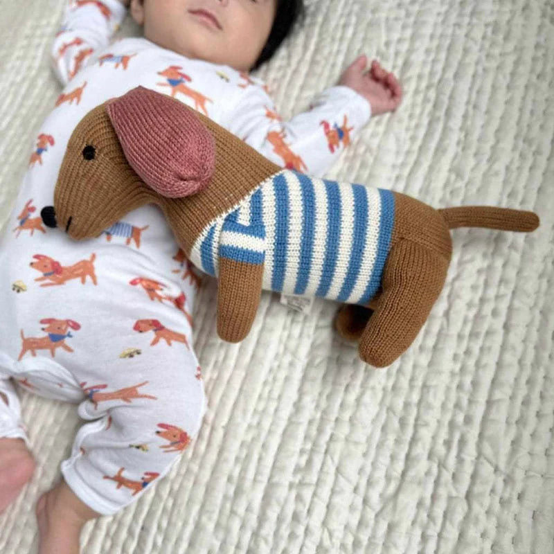 Baby wearing pajamas with dachshund pattern lying next to a knitted dachshund toy on a textured surface.